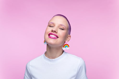 Cheerful young woman wearing white t-shirt and funny rainbow earrings smiling at camera. Studio port...