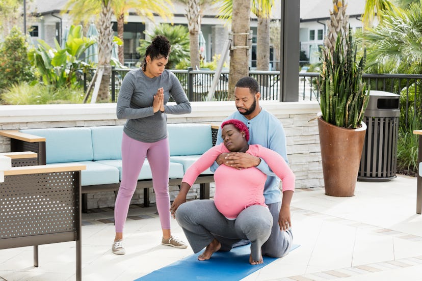 A pregnant Black woman squats in a sunny patio, her Black male partner kneels behind her and is brac...
