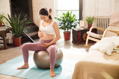 A pregnant Asian woman in a white sports bra and mauve leggings, sits in squatting position on a sil...