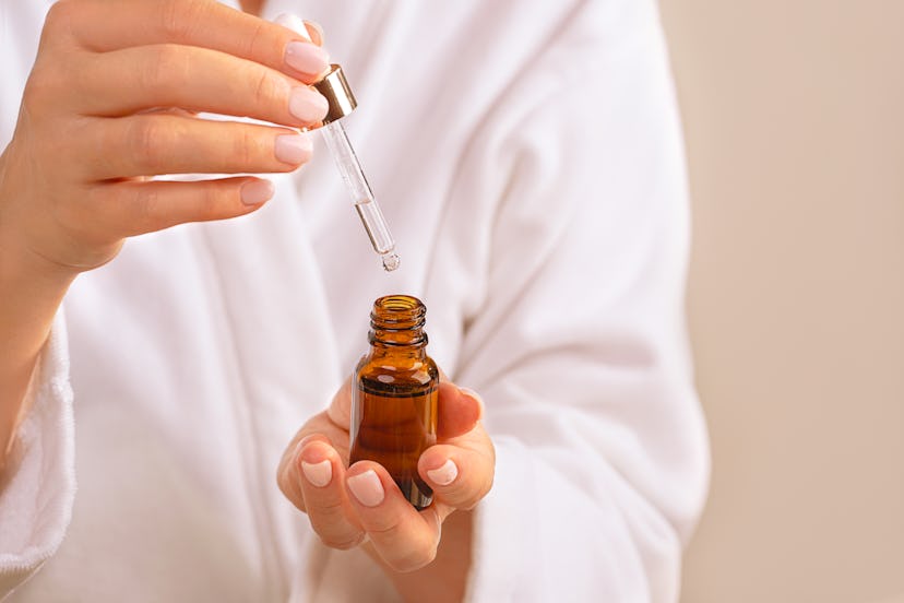 Woman in white bathrobe holds Beauty Product Facial Serum or Essential Oil on trendy 2022 Gray North...