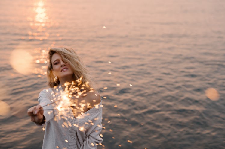 Young woman holding a sparkler at the beach, feeling enchanted by the March 21, 2022 weekly horoscop...