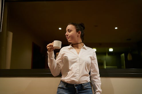 Female wearing a bun and holding a cup of coffee against a glass window in a hotel room