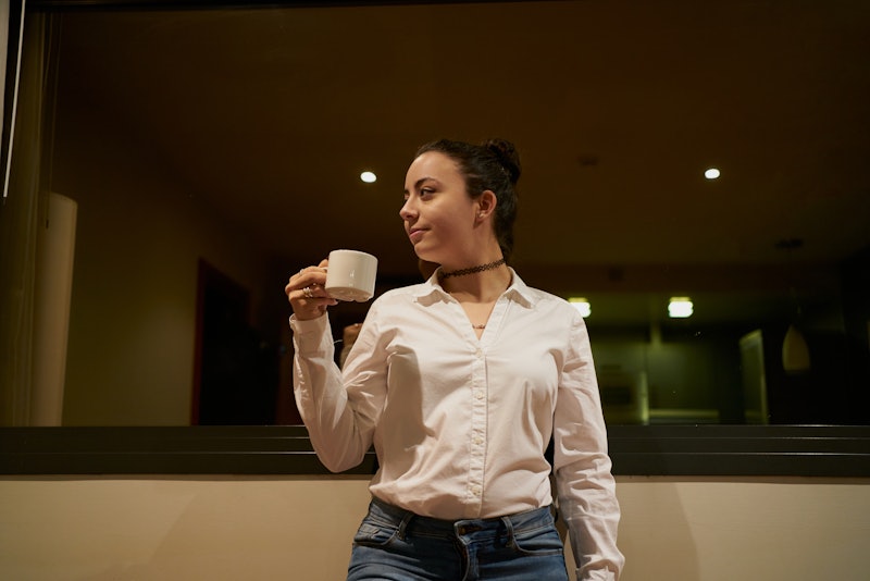 Female wearing a bun and holding a cup of coffee against a glass window in a hotel room