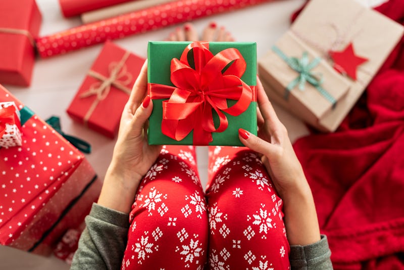 Young woman wearing xmas pajamas sitting on the floor amongst wrapped christmas presents, holding be...