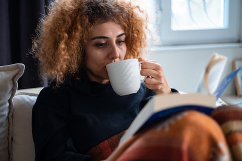 Young woman sitting on sofa with mug of coffee in reading a book.