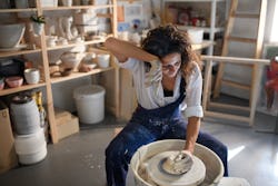 A woman wipes sweat off her face while throwing pottery on the wheel. Here's your daily horoscope fo...