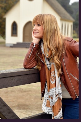 Jane Seymour posing in a brown leather jacket while leaning on a fence with a chapel in the backgrou...