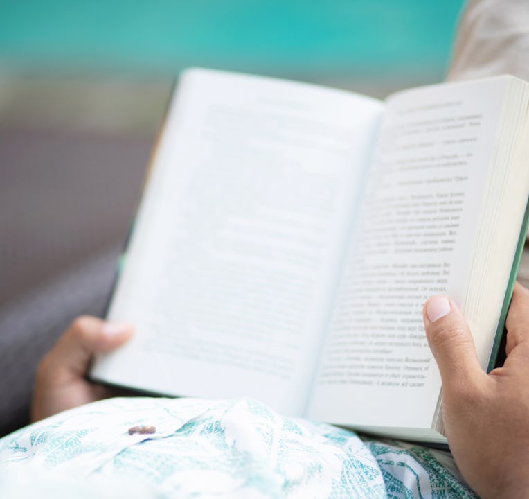 Back view of young man in casual clothes opening and reading a book and relaxing while lying on couc...