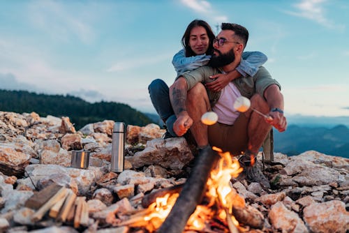 Young couple sitting at the top of the mountain over a fire and roasting marshmallows. These are the...