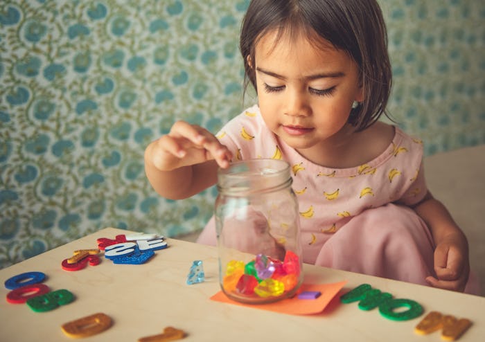 A cute toddler girl learns to count brightly colored toy gems by putting them into a jar one at a ti...