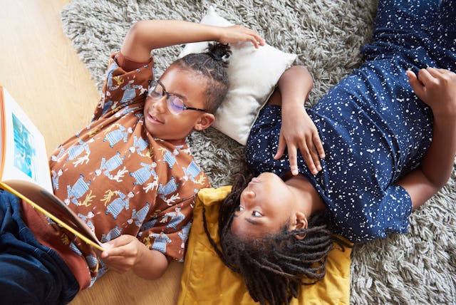 Kids laying on the floor, one is reading a book and the other is listening to him
