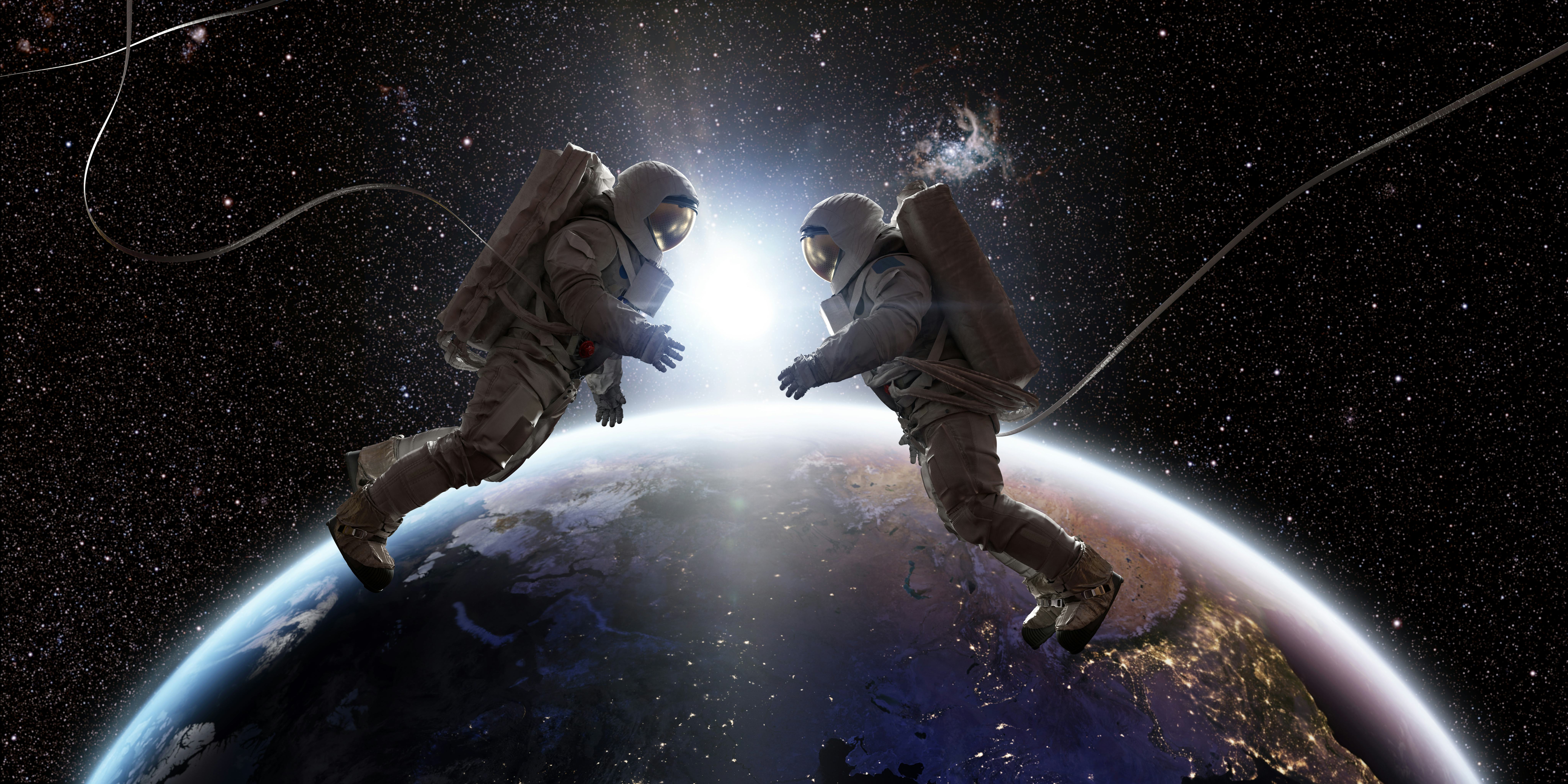 Two astronauts in full spacesuits with backpacks on a space walk with tethers, facing each other wit...