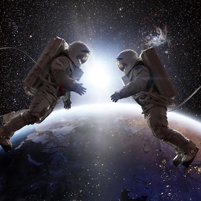Two astronauts in full spacesuits with backpacks on a space walk with tethers, facing each other wit...