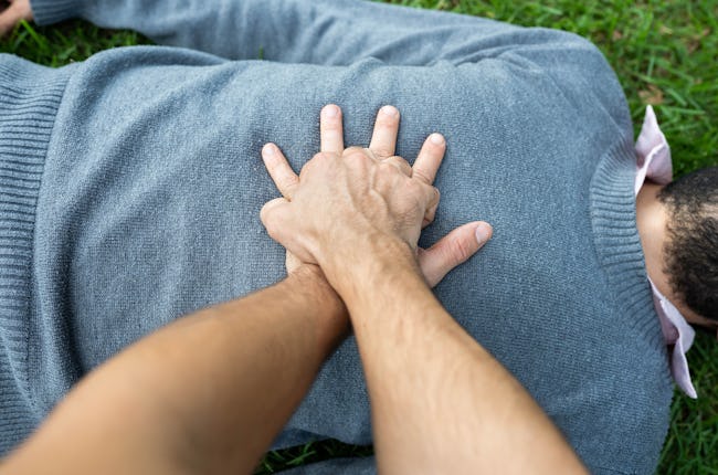A person giving CPR to a black man who is wearing a blue sweater and lying on grass