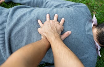 A person giving CPR to a black man who is wearing a blue sweater and lying on grass