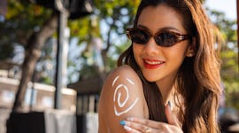 Beautiful happy cute woman applying suntan cream to her shoulder with beach background