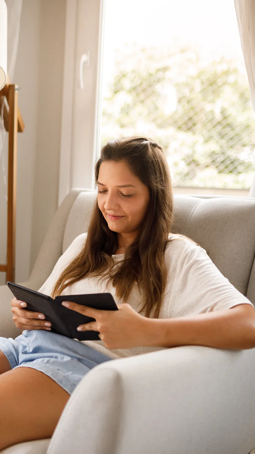 a young woman with long hair sits in a chair, reading an e-book while smiling softly