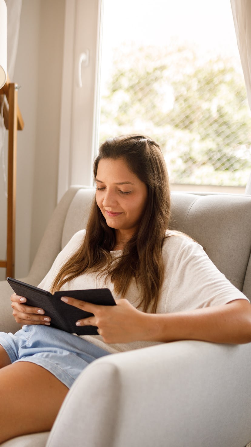 a young woman with long hair sits in a chair, reading an e-book while smiling softly