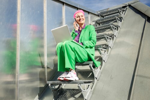 A woman with pink hair and a green suit takes a phone call on her laptop while sitting on stairs. Th...
