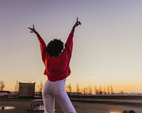 Excited young woman with arms raised standing on roof during dusk. Here's the spiritual meaning of A...