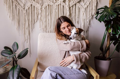 A woman cuddles her dog in front of a macrame hanging. Here's your daily horoscope for March 31, 202...