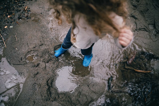 A cute 3 year old Caucasian girl wearing blue rubber boots stomps and splashes in puddles of water n...