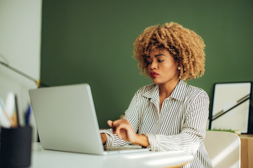 Serious African American woman typing business report on a laptop keyboard while sitting at office d...