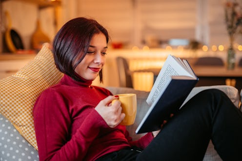 Young beautiful woman lying on sofa at home, drinking coffee and reading book in the evening.