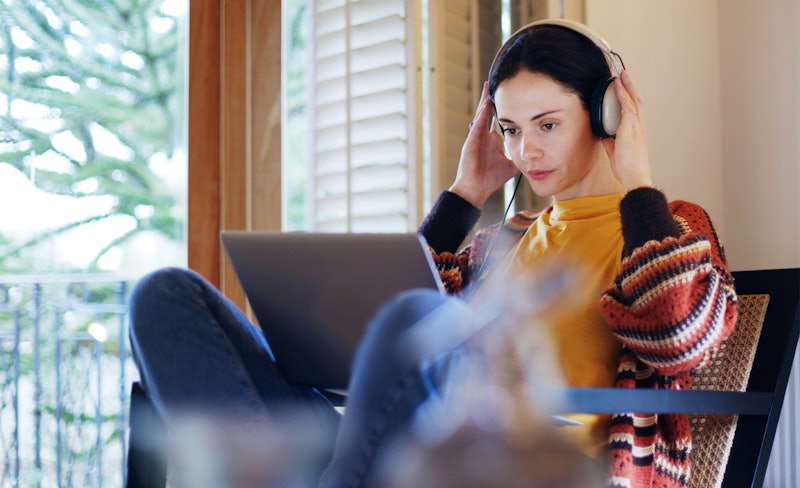 Stock image of a dark-haired young woman reclining at home with her laptop & headphones,  she’s work...
