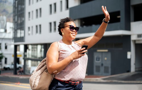 Smiling woman standing on a city street and waving at her taxi pickup. . Aries, Leo, Scorpio, and Ca...