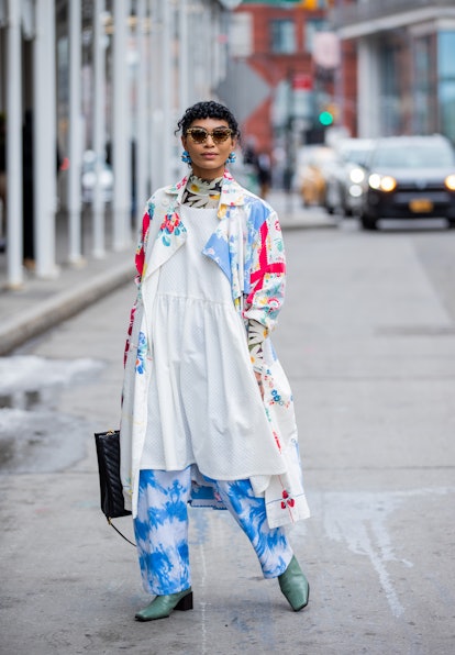 NEW YORK, NEW YORK - FEBRUARY 16: A guest wearing white dress, trench coat with batik print, pants, ...