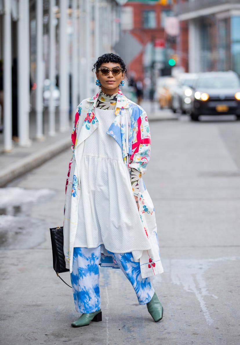 NEW YORK, NEW YORK - FEBRUARY 16: A guest wearing white dress, trench coat with batik print, pants, ...