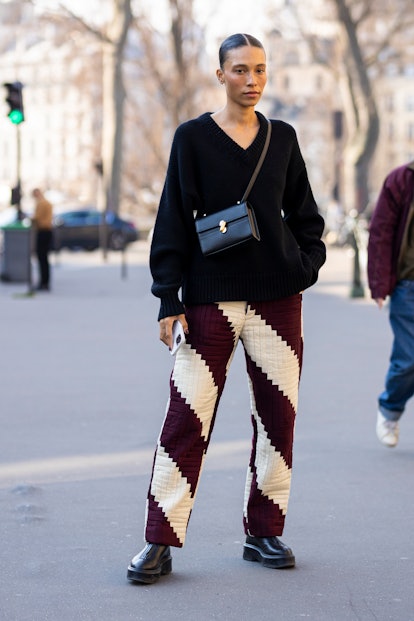 PARIS, FRANCE - MARCH 07: A guest wearing a black sweater, black bag, burgundy and cream pants and b...