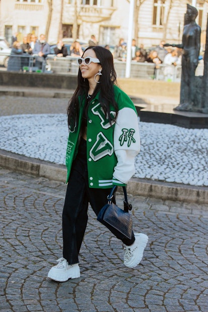 PARIS, FRANCE - MARCH 08: A guest wears a green varsity Louis Vuitton jacket, black pants, black pur...