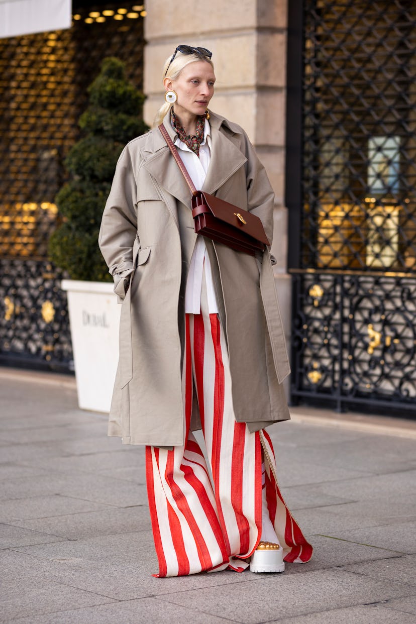 PARIS, FRANCE - MARCH 06: A guest wearing taupe trench coat, an orange and white striped suit, white...