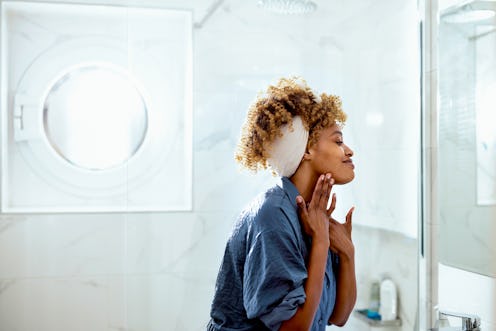Beautiful woman standing in her bathroom having her morning ritual. She is looking at the mirror whi...