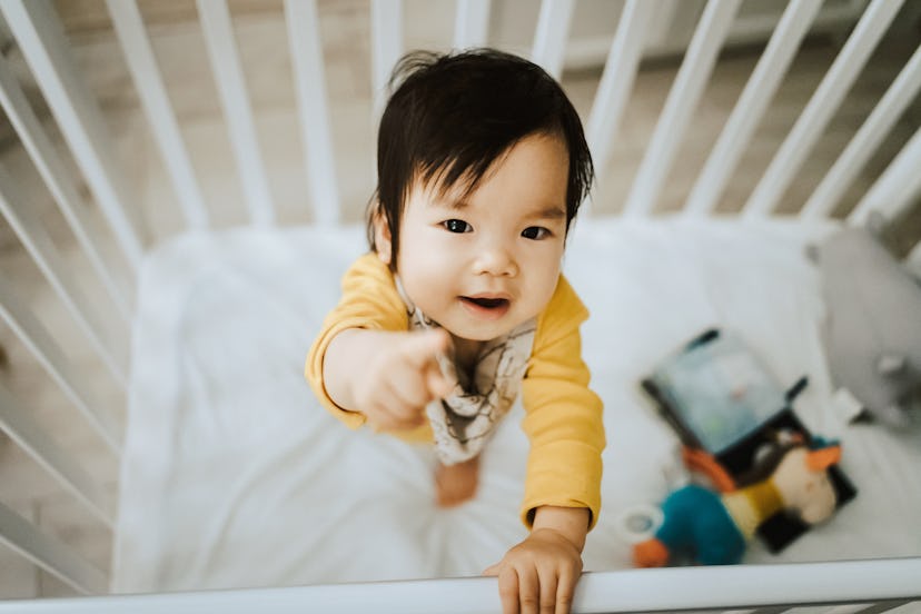 Adorable Asian baby girl standing in her crib and pointing away with fingers