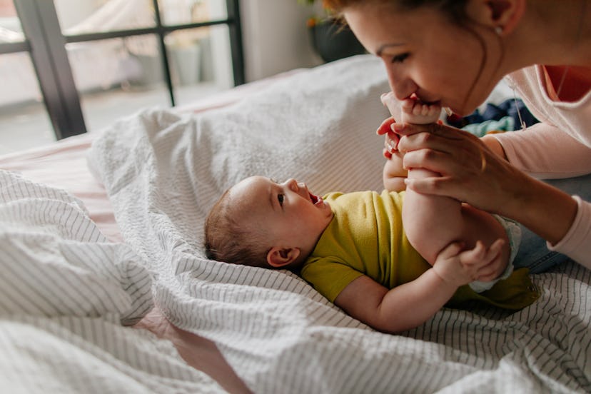Portrait of a little smiling baby boy and his mom, kissing his tiny feet right after waking up in hi...
