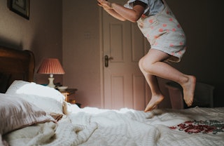 Little girl, in a dated room, leaping and jumping on an old fashioned bed. She is illuminated by sun...