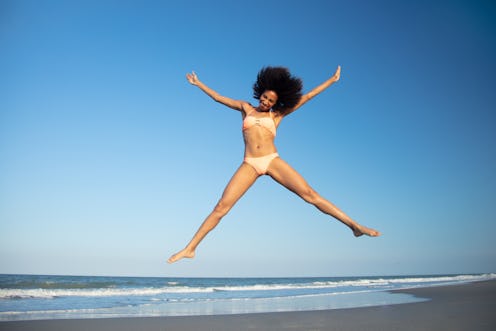 Energetic young Afro American woman jumping high in swimwear and having fun by the ocean on Summer v...