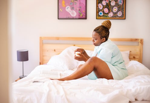 Young african woman sitting on bed and opening a bottle cream for feet in the morning