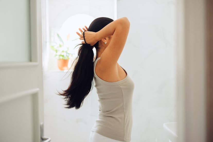 Beautiful Asian woman standing in her bathroom tying her hair  and looking herself in the mirror.