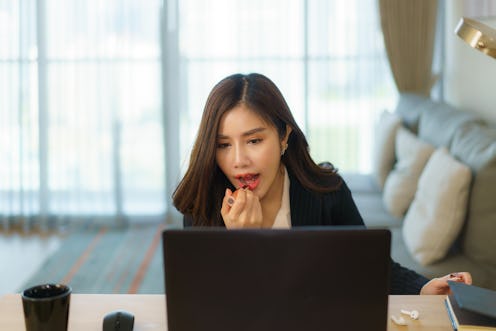 Asian woman getting dressed and putting lipstick on her mouth while preparing for an online meeting ...