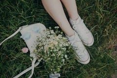 Female legs in white sneakers, bunch of summer daisy flowers, white bag and blue sunglasses on green...