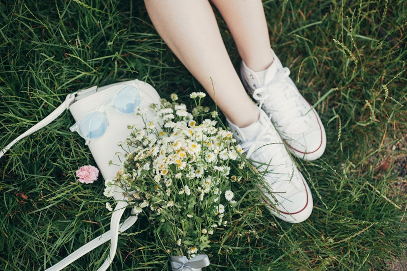 Female legs in white sneakers, bunch of summer daisy flowers, white bag and blue sunglasses on green...