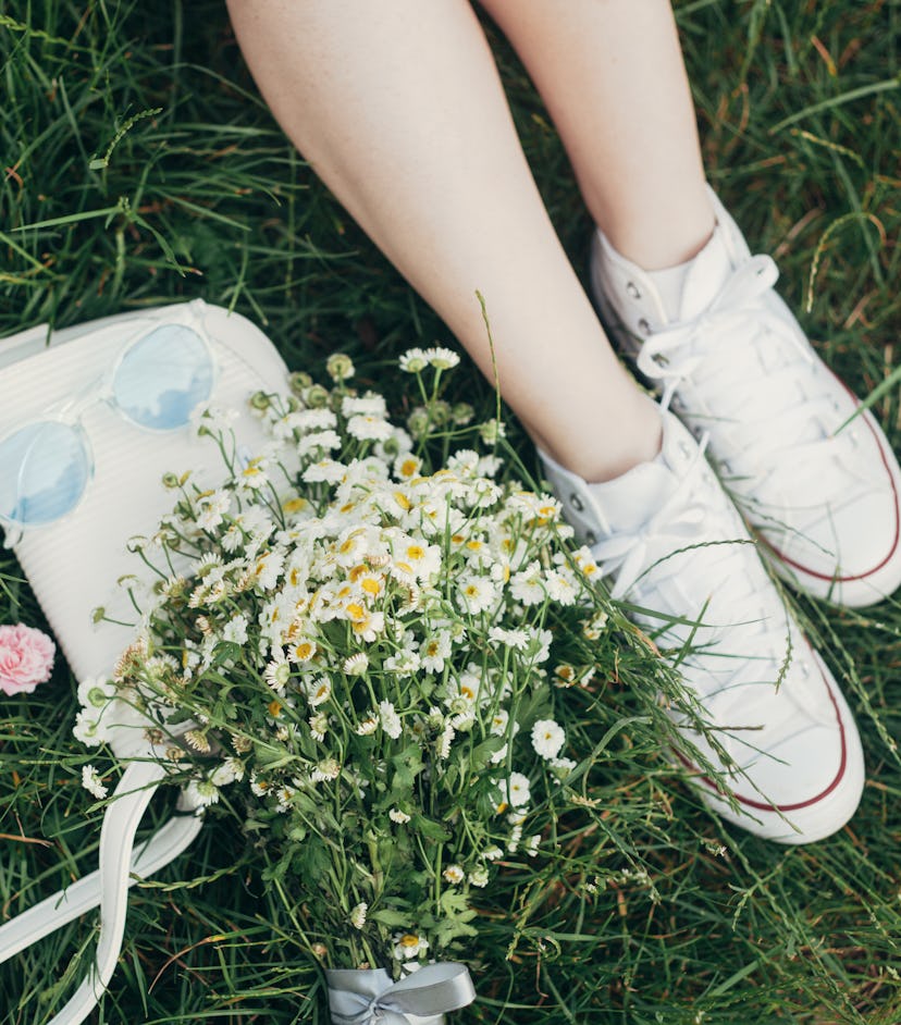 Female legs in white sneakers, bunch of summer daisy flowers, white bag and blue sunglasses on green...