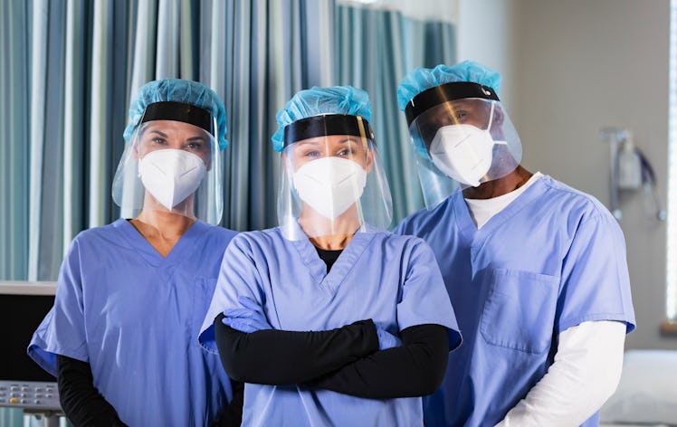 A multiracial group of three healthcare workers, wearing N95 masks and face shields, standing togeth...