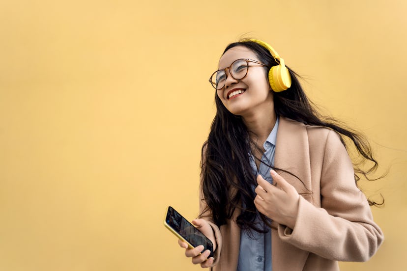 Young asian woman in coat with yellow headphones and mobile phone listening to music via bluetooth a...