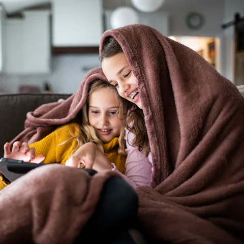 Tween sisters snuggled under blanket and watching a movie on digital tablet.