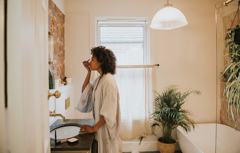 A beautiful black woman stands in a stylish bathroom in a long, silky robe and carefully applies her...